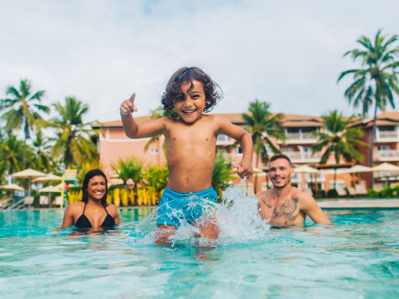 Criança correndo na piscina em direção à camêra, dentro da piscina da Costa do Sauípe, com os pais ao fundo