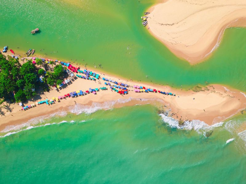 Visão aérea de uma das pequenas ilhas de Caraíva, cercada de mar esverdeado e várias cadeiras e barracas coloridas na areia