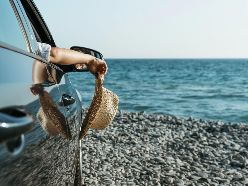 Parte de um carro elétrico parado de frente ao mar, com uma pessoa com a braço pra fora segurando um chapéu de praia